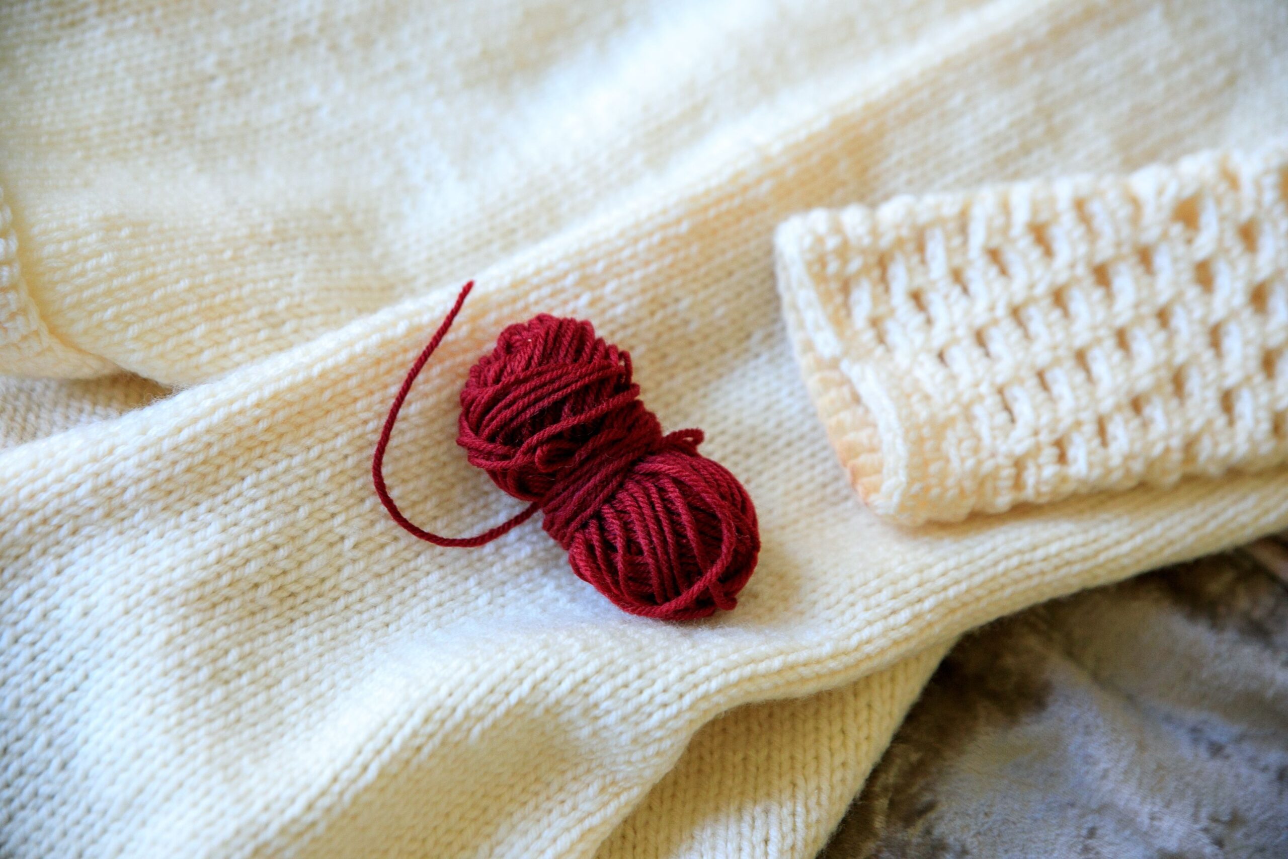 A ball of deep red yarn resting on cream knitted wool, the only saturated colour in the frame