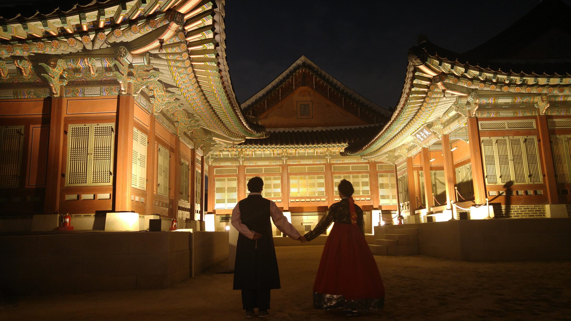 A couple in hanbok walking through a lit Gyeongbokgung courtyard during the night opening