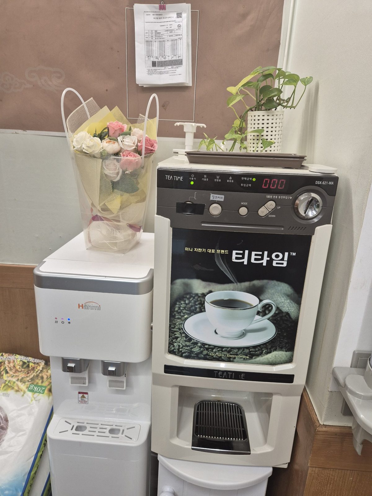 A small Tea Time coffee vending machine with a coin slot and a 'milk coffee' button, next to a water dispenser, with a pork grading certificate clipped to the wall above.