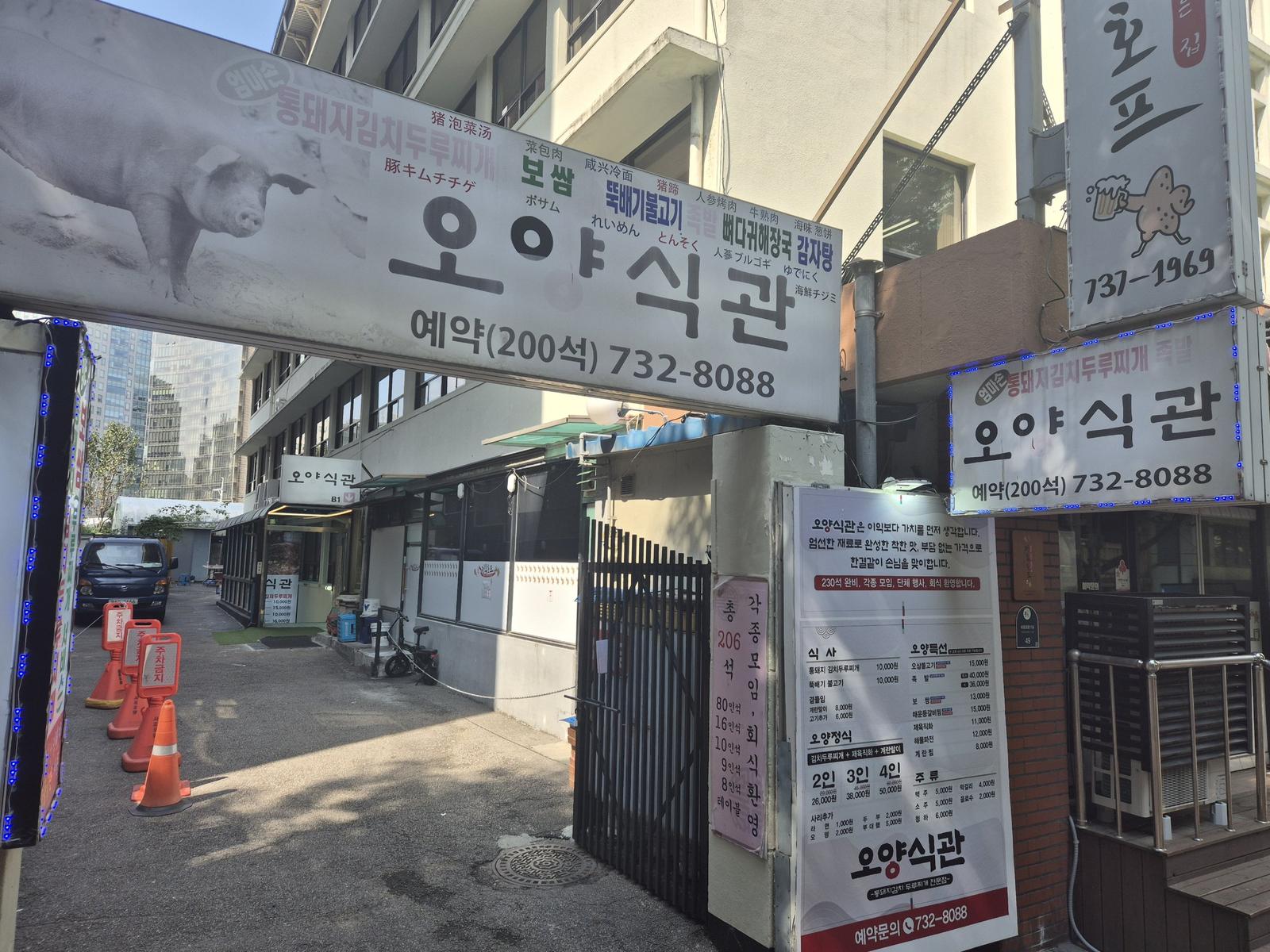 The entrance to Oyang Sikgwan off Sejong-daero 21-gil: a long white sign with a photograph of a pig, the restaurant name, a list of dishes in Korean, Chinese and Japanese, and the reservation number 732-8088 above a short driveway with parking cones.