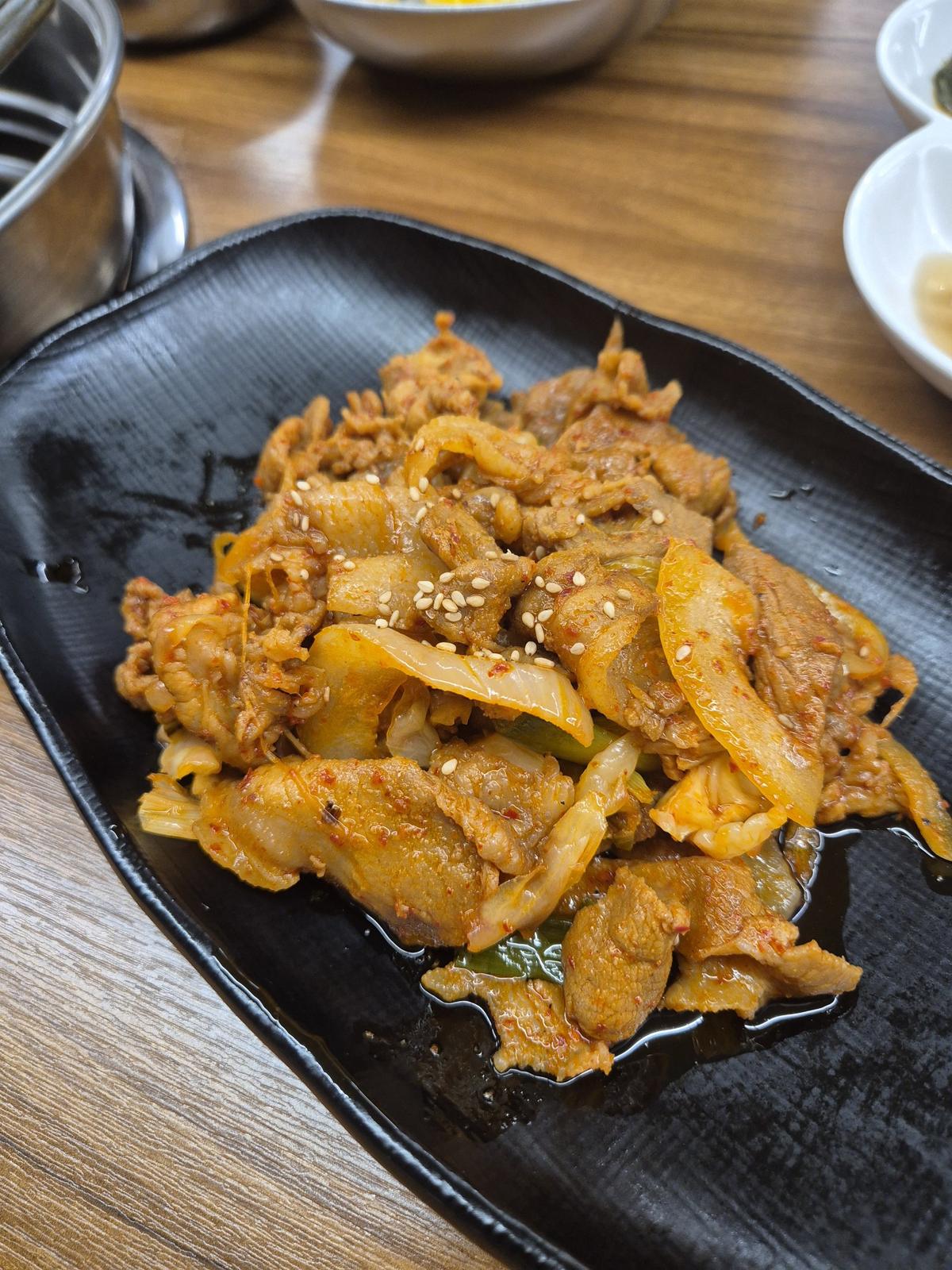 A black plate piled with spicy stir-fried pork, onion and spring onion in a glossy red chilli sauce, scattered with sesame seeds.