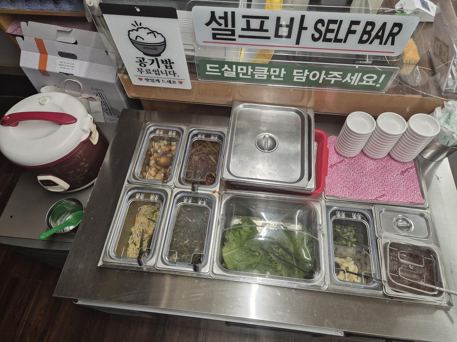 A stainless-steel self-service bar with tubs of pickled radish, sweet pickled chillies, bean sprouts, seasoned greens, lettuce, green chillies, garlic and ssamjang, a rice cooker on the left and stacks of small dishes on the right.