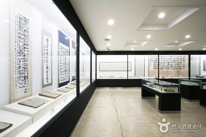 A bright exhibition room with display cases of old books and tall hanging scrolls of Hangeul calligraphy.