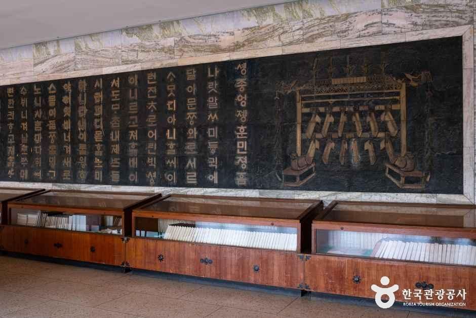 A long black wall carved with the opening lines of Hunminjeongeum in gilt Hangeul beside a relief of a bell frame, with glass cases of books below.