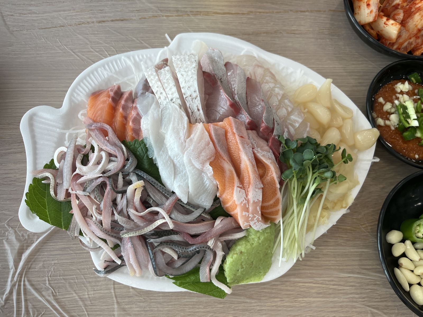 A mixed sashimi platter on a fish-shaped plate: thin silver slices of gizzard shad at bottom left, white flounder in the middle, sea bream with its skin pattern and silver-skinned jack at the top, clear sea bass on the right and orange salmon, with pickled rakkyo, radish sprouts and wasabi alongside.
