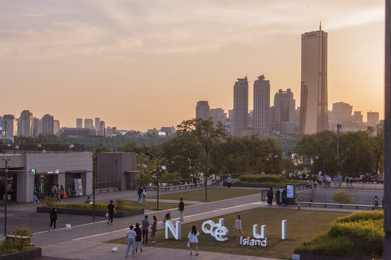 The 'Nodeul Island' letter sculpture and lawn on the island's plaza, people walking past, with the 63 Building and Yeouido towers in the sunset behind.