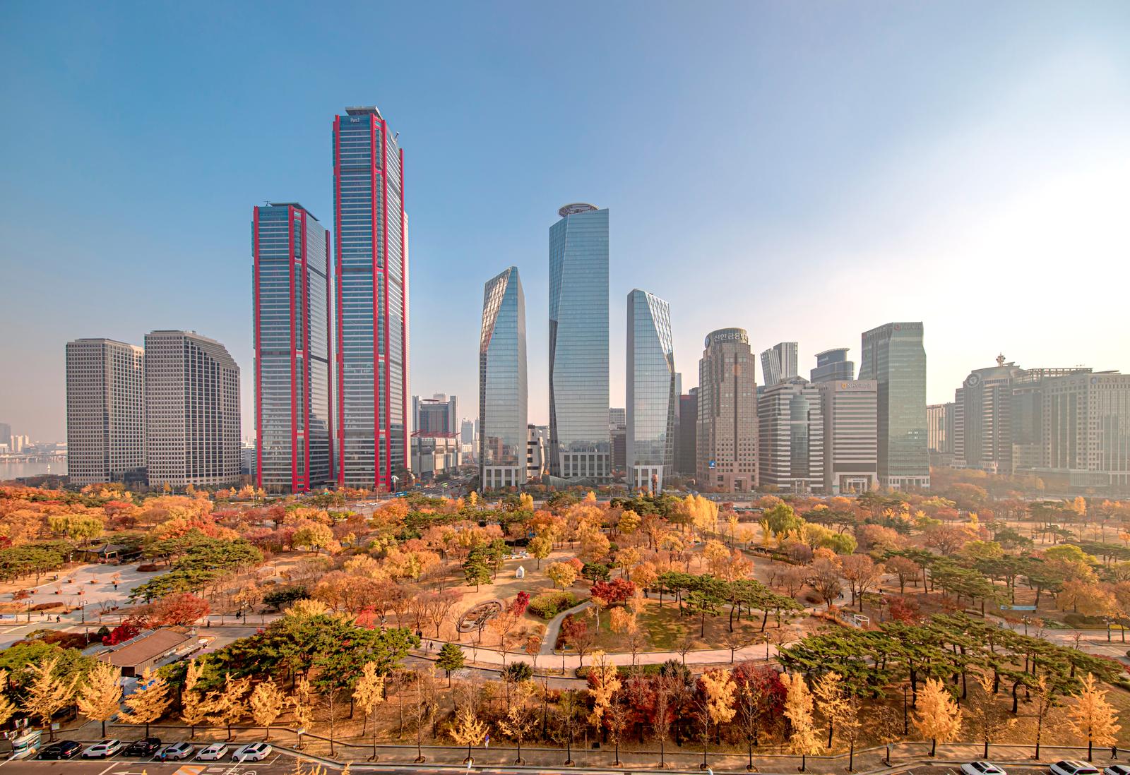 Yeouido Park in autumn: trees turned yellow and red in front of a red-framed tower and glass office buildings.